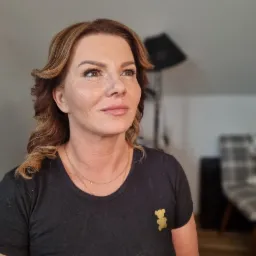 Portret kobiety z delikatnym makijażem, podkreślonymi brwiami i ustami w odcieniu nude, włosy w odcieniach brązu i blondu, z lokami opadającymi na ramiona, ubrana w czarną koszulkę z aplikacją...